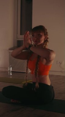 Dikey ekran: Turuncu spor sutyenli bir kadının, loş ışıklandırılmış stüdyoda yoga dersi sırasında dua pozisyonunda meditasyon yapması. Farkındalık ve iç huzur kavramı.