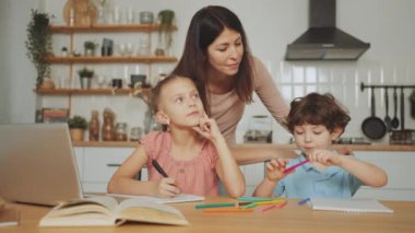 Anne masada iki çocuğa ödevlerinde yardım ediyor. Bir çocuk deftere yazarken, diğeri pastel boyalarla oynuyor. Evde eğitim ve ebeveynlik kavramı. Evde öğrenim ve eğitim