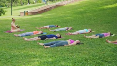 Yoga sınıfı katılımcıları, parkta şarkı söyleyen bir eğitmenin rehberliğinde meditasyonda yatıyor. Rahatlamaya, uyuma ve farkındalığa odaklan. Dinginlik ve kolektif enerji kavramı