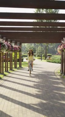 Parkta ahşap pergola altında mavi bisiklete binen kadın. Sarı kıyafetli ve hasır şapkalı. Güneşli bir günde etrafım çiçekler ve yeşilliklerle çevrili. Açık hava aktiviteleri ve boş zaman kavramları.