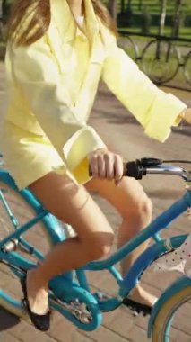 Dikey ekran: Sarı elbiseli mavi bisiklete binen kadının yakın çekimi. Ellerini gidona koy, güneşli günün tadını çıkar. Açık hava aktiviteleri ve sağlıklı yaşam tarzı kavramı