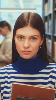 Dikey ekran: Kütüphane koridorunda elinde defterlerle kameraya bakan ciddi bir öğrenci. Renkli kitaplar ve başka bir öğrenci okuması. Odaklanmış öğrenme ve akademik adanmışlık kavramı