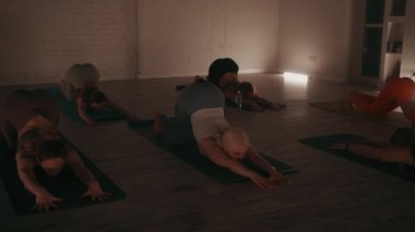 Bir grup kadın, loş ışıklı stüdyoda yoga minderleri üzerinde uzun köpek pozu pratiği yapıyor. Ilık aydınlatma ve aynalar huzurlu bir atmosfer yaratır. İç huzur ve esneklik kavramı