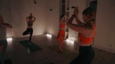 Bir grup kadın, loş ışıklı stüdyoda yoga minderlerinde ağaç pozu veriyor. Katılımcılar spor sutyen ve tayt giyerler. Ilık ışıklandırma sakin bir atmosfer yaratır. Denge ve konsantrasyon kavramı