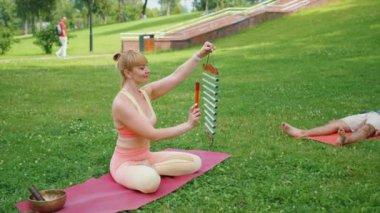 Yoga eğitmeni, grup meditasyonunda açık hava yoga seansı sırasında rüzgar çanları kullanıyor. Paspaslara uzanan katılımcılar, huzur parkındaki rahatlama ve farkındalığa odaklanıyorlar. Ses iyileştirme kavramı