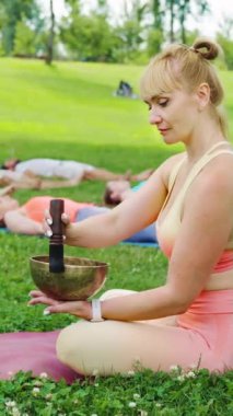 Yoga eğitmeni, grup meditasyonunu açık hava yoga seansı sırasında şarkı kasesi kullanarak yapıyor. Katılımcılar paspaslara uzanıp, huzur parkındaki rahatlama ve farkındalığa odaklanıyorlar.