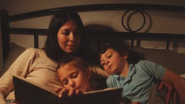 Anne ve iki çocuğu yatakta kucaklaşırken kız da masal okuyor. Aile bağları ve uyku zamanı ritüelleri kavramı. Sevgiye, rahatlığa ve çevreye odaklan