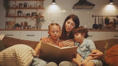 Anne ve iki çocuğu kanepede otururken kızı yüksek sesle kitap okuyor. Aile bağı ve okuma sevgisi kavramı. Evde öğrenme ve duygusal bağ geliştirme