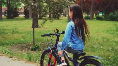Kendine güvenen kız parkta bisiklet sürüyor, tek eliyle gidonu tutuyor. Kot ceket giyip açık hava aktivitelerinin tadını çıkarıyorum. Çocukluk güveni, özgürlük, bağımsızlık ve macera kavramı
