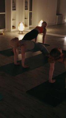 Dikey ekran: Bir grup kadın, loş ışıklandırılmış stüdyoda yoga minderleri üzerinde yukarı doğru tahta pozu veriyor. Ilık aydınlatma ve aynalar sakin atmosferi güçlendirir. Güç ve esneklik kavramı.