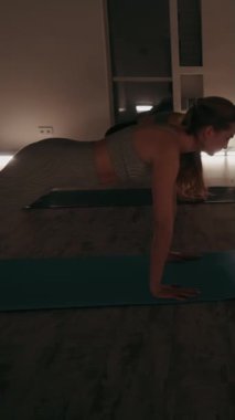 Dikey ekran: Bir grup kadın, loş ışıklandırılmış stüdyodaki yoga minderleri üzerinde aşağı doğru köpek pozu sahnesine alıştırma yapıyor. Ilık ışık, sakin bir atmosfer yaratır. Güç ve esneklik kavramı.