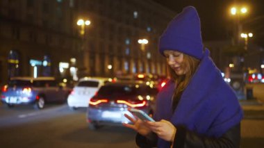 Mavi şapkalı ve atkılı genç bir kadın elinde akıllı telefon tutarken trafiğe bakıyor. Kadın gece şehir caddesinde duruyor. Arka planda bulanık arabalar ve ışıklar var. Kentsel yaşam tarzı konsepti