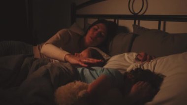 Anne, uyuyan iki çocuğuyla yatağa kıvrılıp huzurlu ve sıcak bir atmosfer yaratıyor. Uyku zamanı rutini ve aile bağları kavramı. Aile hayatındaki rahatlığa ve birlikteliğe odaklan.