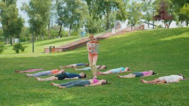 Yoga eğitmeni, açık hava oturumunda rüzgar çanları kullanarak katılımcılara son rahatlama boyunca rehberlik ediyor. Katılımcılar derin bir rahatlama için parktaki paspasların üzerine yatıyorlar. Farkındalık ve meditasyon kavramı