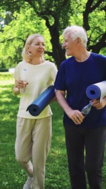 Dikey ekran: Parkta yürürken yoga minderleri taşıyan ve su şişeleri tutan yaşlı çift. Kıdemli fitness, hidrasyon, sağlıklı yaşam tarzı kavramı