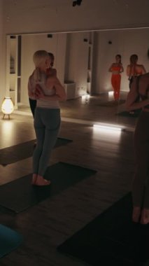 Dikey ekran: Bir grup kadın, loş ışıklandırılmış stüdyoda yoga minderleri üzerinde elleri arkadan kenetlenmiş şekilde sırt bükme pozu veriyor. Esneklik ve denge kavramı