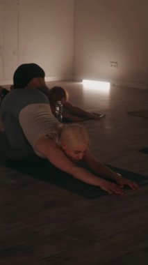 Dikey ekran: Katılımcılar loş ışıklı stüdyoda yoga seansına katılıyorlar, esneme ve gevşemeye odaklanıyorlar, paspaslar üzerinde çocuk pozu veriyorlar, dingin ve rahatlatıcı bir atmosfer yaratıyorlar.