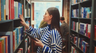Çizgili süveter giyen öğrenci kütüphane koridorunda kitap arıyor, elinde tablet var. Geçmişi renkli kitaplarla dolu ve başka bir öğrenci daha okuyor. Odaklanmış araştırma ve akademik keşif kavramı