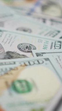 Dikey ekran: Masaya dağılmış yüz dolarlık banknotlar. Amerika 'nın yüz dolarlık banknotlarıyla arka planda dönüyor. İş ve finans kavramı