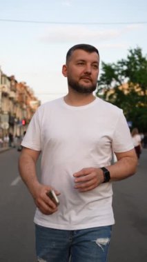 Beyaz tişörtlü ve kot pantolonlu rahat sakallı adam şehrin hareketli caddesinde yürüyor, telefon elinde, alacakaranlıkta tarihi binaların önünde. Her gün şehir hayatı