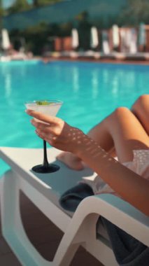 Kadın havuz kenarında dinleniyor, serinletici bir kokteyl tutuyor. Güneşin altında rahatlama, rahatlama ve keyif. Yaz tatili kavramı, havuz kenarındaki lüks ve kaygısız anlar.