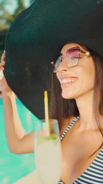 Siyah güneş şapkası ve güneş gözlüğü takan, elinde bilardo içen gülümseyen bir kadının yakın çekimi. Yaz neşesi, rahatlama ve lüks kavramı