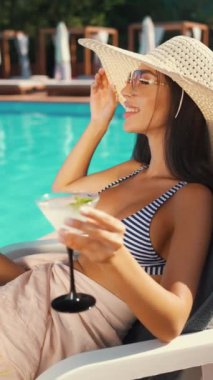 Kadın havuzda rahatlıyor, gülümsüyor ve güneş şapkası ve çizgili bikini giyerken ferahlatıcı bir kokteyl tutuyor. Yaz tatili, rahatlama ve keyif. Tatil kavramı, lüks ve tasasız anlar.