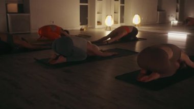 Bir grup çocuklu kadın yoga minderlerine poz veriyor. Sakin, loş bir stüdyoda. Çeşitli renkli spor sutyenler ve taytlar. Sıcak ortam ışıkları ve aynalar sakin atmosferi geliştirir. Rahatlama kavramı