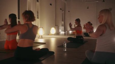 Yoga eğitmeni. Huzurlu ve loş bir stüdyoda kadın sınıfının lideri. Oturan katılımcılar dua pozisyonu almış şekilde poz veriyorlar. Sıcak ortam aydınlatma ve aynaları sakin atmosferi geliştirir.