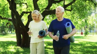 Yaşlı çift parkta egzersiz yaptıktan sonra yoga minderleri tutarak su içiyor. Yeşil çevre sağlıklı bir yaşam tarzını vurgular. Zindelik, sağlık ve aktif yaşlanma kavramı.