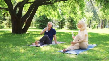 Yaşlı çift parkta yoga minderlerinde oturuyor, su içiyor ve antrenmandan sonra dinleniyor, sonraki yıllarda aktif kalıyorlar. Etkin yaşlanma, sağlık ve susuzluk kavramı