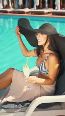 Kadın bilardo salonunda, elinde ferahlatıcı bir içecek tutarken büyük siyah güneş şapkası ve çizgili bikini giyiyor. Tatil kavramı, lüks ve tasasız rahatlama.