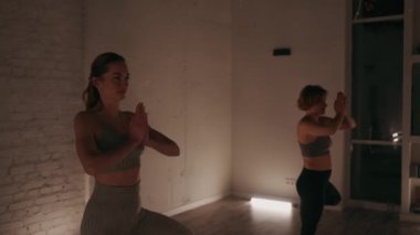 Bir grup kadın, loş ışıklı stüdyoda yoga minderlerinde ağaç pozu veriyor. Ilık aydınlatma ve aynalar huzurlu bir atmosfer yaratır. Denge ve konsantrasyon kavramı