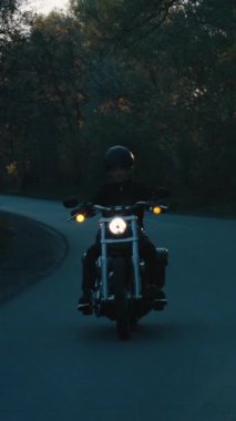 Dikey ekran: Siyah elbiseli bisikletçi ve kask takan motosikletli yavaş çekimde kır yolunda motosiklet süren, güneşin ağaçların arasından geçmesini sağlayan. Mistik kasvetli doğa. Macera kavramı