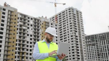 Yansıtıcı yelek ve miğfer mühendisi inşaat alanında dizüstü bilgisayarla çalışıyor. Yüksek binalar ve arka planda inşaat vinci. Modern inşaat yönetimi kavramı