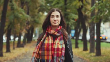 Uzun saçlı ve ekose eşarplı kadın sonbahar parkında huzurlu bir gezinti yapıyor. Etrafı canlı sonbahar yaprakları ve kentsel geçmişle çevrili. Özgüven ve mevsimsel güzellik kavramı