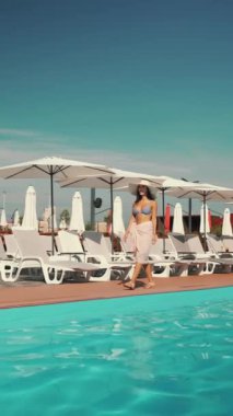 Gülümseyen kadın, açık pembe pareo 'yu çıkarırken çizgili bikini ve güneş şapkasıyla havuz kenarında geziniyor. Beyaz şezlonglar ve şemsiyeler arka planda. Yaz tatili ve zarafet kavramı.
