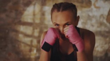 Saçlarını boks pozisyonunda örmüş, pembe el sargıları takan kararlı bir kadın. Yoğunluğu yakalar ve kumlu spor salonlarına odaklanır. Yetki ve disiplin kavramı