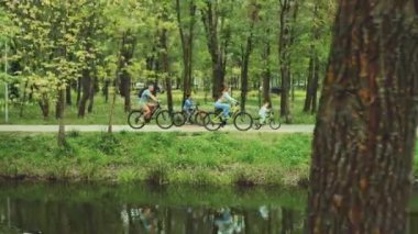 Dört kişilik bir aile gölet boyunca yeşil parktan bisikletle geçiyorlar. Ebeveynler ve çocuklar açık hava aktivitelerini ve temiz havayı severler. Kavram: Aile bağı, sağlıklı yaşam tarzı, doğa bağlantısı
