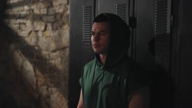 Kapüşonlu bir adam elleri yüzü asılmış, kırmızı sargılar takmış, gözleri tükenmiş bir şekilde dolaplara yaslanıyor. Loş ışık. Yorgunluğu ve azmi kavra
