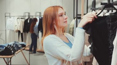 Moda mağazasında giysileri dikkatle inceleyen, alışveriş deneyimini temsil eden ve yeni kıyafetler seçen genç bir kadın. Moda anlayışı, perakende terapisi ve kişisel tarz.