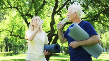 Yaşlı adam ve kadın yeşil parkta yoga minderleri tutarken şişelerden su içiyorlar. Fiziksel aktiviteden sonra ferahlatıcı
