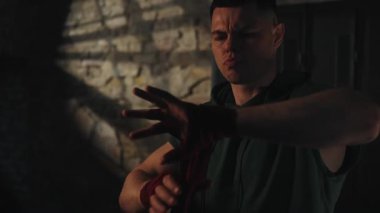 Kolsuz ve kırmızı kapüşonlu bir adam, duvarları tuğla kaplı loş spor salonundaki yumruklarıyla bandajları sıkıyor ve yoğun egzersize hazırlanıyor. Kavram: disiplin, hazırlık