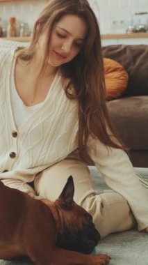 Dikey Ekran: Genç bir kadın yerde oturmuş sıcak oturma odasında Fransız Bulldog 'la oynuyor. Hayvan bakımı ve bağlanma kavramı. Kız nazikçe köpeğini okşuyor. İçerde sevgi dolu bir an yaratıyor.