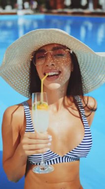 Çizgili bikinili ve geniş hasır şapkalı kadın elinde samanla bir bardak limonata tutarken ahşap havuz güvertesinde oturmuş parlak mavi su yüzeyinin yakınında gülümsüyor.