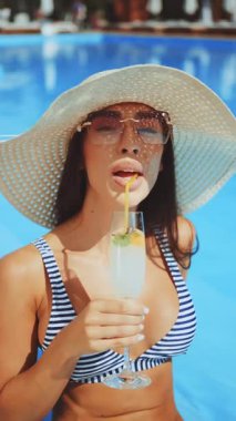 Çizgili bikinili ve büyük hasır şapkalı gülümseyen genç kadın havuz kenarında güneşli ve açık mavi gökyüzü altında otururken limon dilimi ve samanla kokteyl tutuyor.