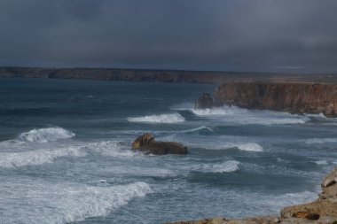 Okyanus manzarası ve kayalık kıyı önlerinde kayalıklar ve dalgalar var.