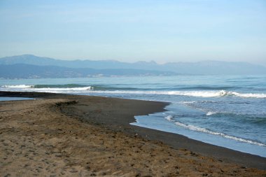 Kumlu kumlu sahil, dalgalar savruluyor, arka planda dağ, mavi gökyüzü, gün sonu ışığı