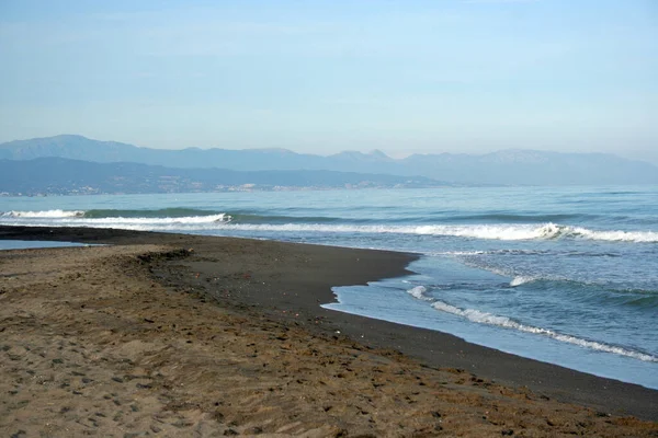 Kumlu kumlu sahil, dalgalar savruluyor, arka planda dağ, mavi gökyüzü, gün sonu ışığı