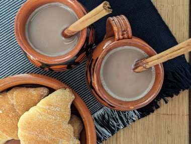 Tarçın çubuklu kil bardakta iki sıcak çikolata, Rattan arka planında el yapımı mavi masa örtüsü üzerinde tatlı Meksika ekmeği.